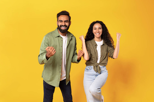 Wow, yes. Joyful arab man and woman cheering and shaking clenched fists over yellow studio background, looking at camera in excitement. Happy couple celebrating win gesturing in joy - Photo, Image
