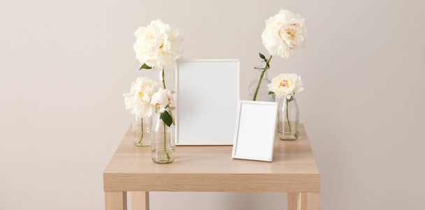 Jarrones con flores de peonía blanca y marcos de fotos en blanco en la mesa de madera cerca de la pared de luz. Banner para el diseño - Foto, imagen