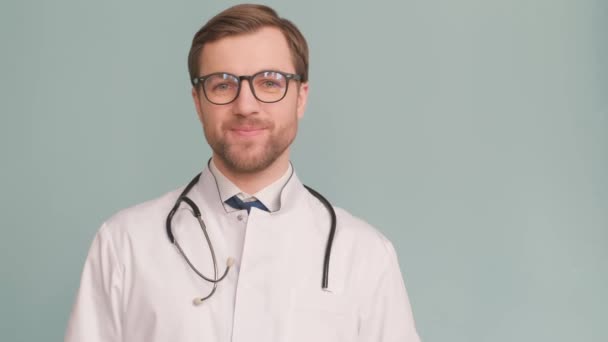 青い背景の上にポーズをとっているヨーロッパの若い医者の笑顔は、驚くほど幸せです。有望な若い医師の広告撮影。医者は親指を立てて - 映像、動画