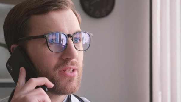 Portret atrakcyjnego lekarza w okularach rozmawiającego przez telefon. Dni pracy lekarza. Profesjonalny młody stażysta - Materiał filmowy, wideo