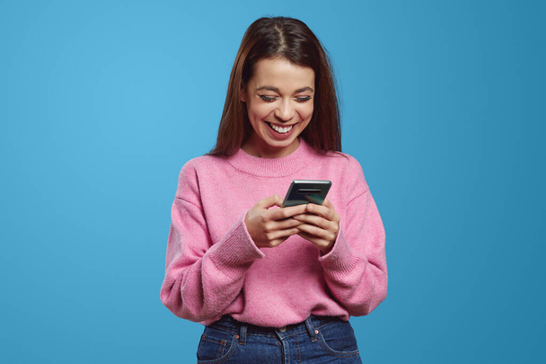 Positivo giovane ispanico femminile millenario sorridente felicemente durante la messaggistica sul telefono cellulare su sfondo blu - Foto, immagini