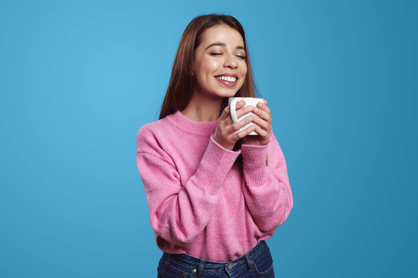 Mujer joven y bonita disfrutando de la bebida con los ojos cerrados y sosteniendo la taza con ambas manos usando suéter rosa, aislado sobre fondo azul - Foto, Imagen