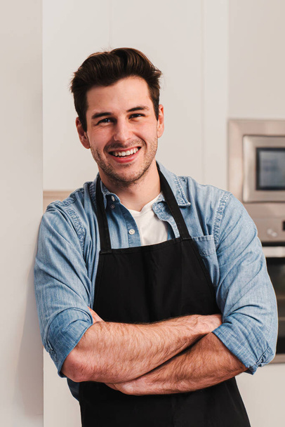 Portrait vertical d'un bel homme caucasien portant un tablier debout dans la cuisine, souriant et croisant les bras regardant la caméra. Vue de face d'heureux beau mâle appuyé contre un mur à la maison - Photo, image