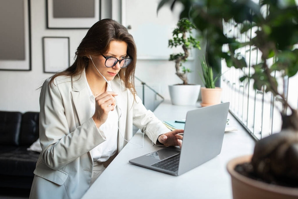 Modern ofis içinde dizüstü bilgisayarda çalışan kulaklıklı neşeli Avrupalı bayan iş toplantısı yapıyor ya da müşteriyle çevrimiçi iletişim kuruyor. Destek ve mali danışmanlık - Fotoğraf, Görsel