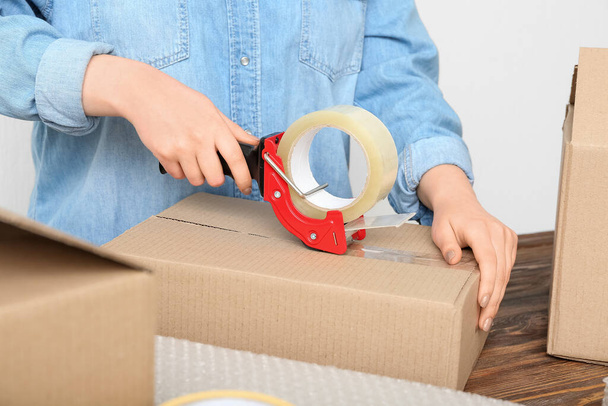 Woman packing cardboard box with adhesive tape dispenser at wooden table - Фото, изображение
