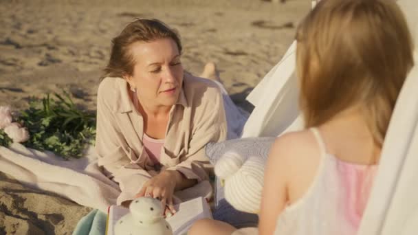 彼女はおしっこテントの下で砂浜に横たわっている間、白人の母親は本を保持し、かわいい女の子におとぎ話を読んでいます.Little blonde kid talking with mother while sitting in cozy tent at sunset 4K - 映像、動画