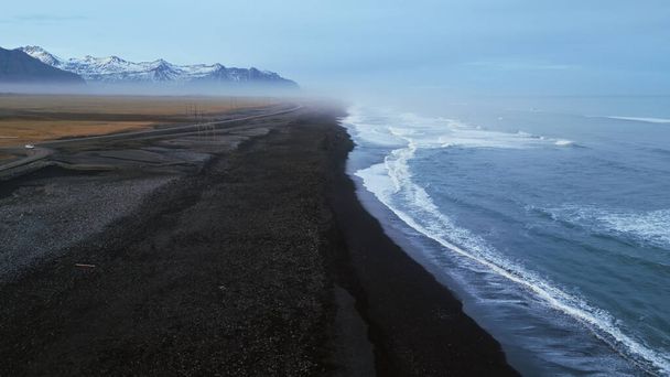 Исландский черный песчаный пляж дрон выстрел, красивая береговая линия с волнами, разбивающимися на атлантическом берегу. Северный ландшафт с заснеженными горами и черным песком на пляжах, живописный маршрут. Медленное движение. - Фото, изображение