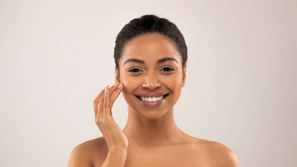 Closeup studio shot of happy pretty young half-naked black woman touching face and smiling, applying nourishing anti-age or hydrating cream, isolated on gray background, panorama - Foto, afbeelding