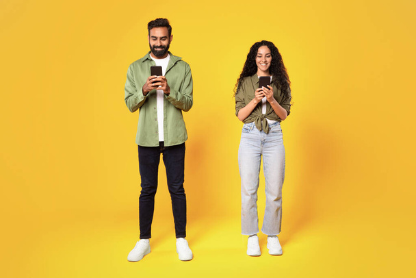 Κινητή επικοινωνία. Χαμογελώντας μεσανατολίτικο ζευγάρι γραπτών μηνυμάτων κρατώντας Smartphones στέκεται σε κίτρινο φόντο στούντιο. Άραβας άνδρας και γυναίκα που χρησιμοποιεί εφαρμογή στα τηλέφωνα. Πλήρης βολή μήκους - Φωτογραφία, εικόνα