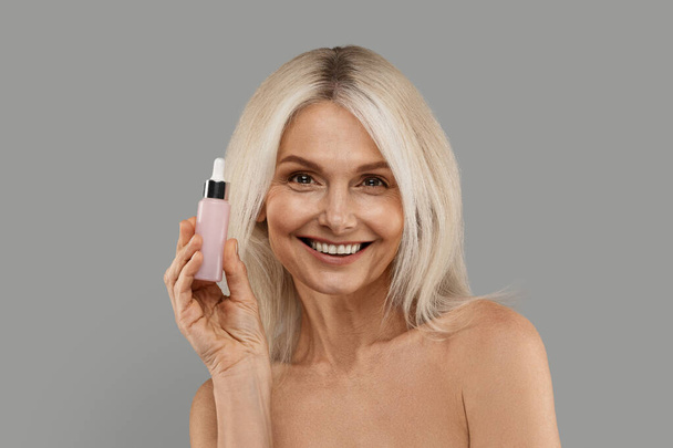 Skincare Concept. Beautiful Mature Female Holding Bottle With Moisturising Face Serum And Smiling, Nude Senior Woman With Beautiful Skin Standing Over Grey Studio Background, Copy Space - Photo, Image