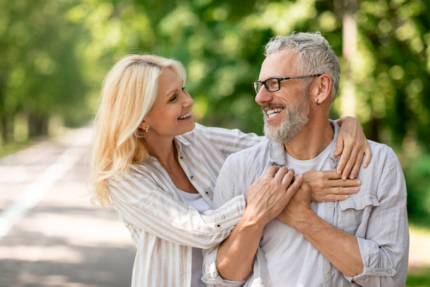 Šťastný romantický dospělý pár objímající při procházce spolu v městském parku, usmívající se starší manžel a manželka objímající a dívající se na sebe, veselý starší manželé užívající si rande venku - Fotografie, Obrázek