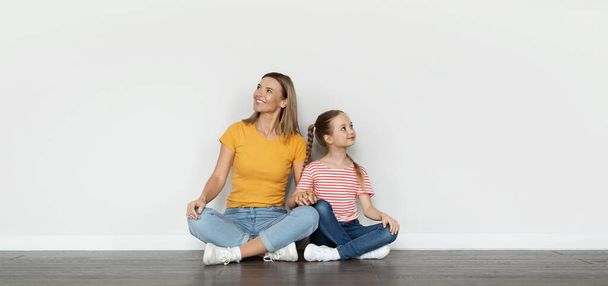Ευτυχισμένη νεαρή μαμά και η μικρή κόρη κοιτάζοντας επάνω στο χώρο αντιγράφων με ενδιαφέρον, μητέρα και χαριτωμένο θηλυκό παιδί κάθονται μαζί στο πάτωμα κοντά στο λευκό τοίχο, Πανόραμα με ελεύθερη θέση για διαφήμιση - Φωτογραφία, εικόνα