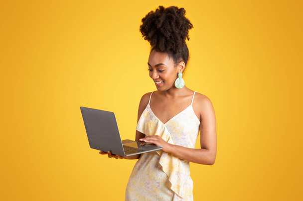 Lachende duizendjarige Afrikaans-Amerikaanse dame typen op laptop, chatten in sociale netwerken, geïsoleerd op gele achtergrond, studio. Werk en studie, reclame en aanbod, apparaat voor freelance - Foto, afbeelding