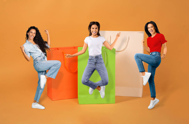 Big Sale. Drei freudige brünette Damen gestikulieren Ja posieren in der Nähe großer Einkaufstaschen auf orangefarbenem Hintergrund. Collage mit riesigen bunten Papiereinkäufern und aufgeregten Shopaholics - Foto, Bild