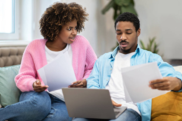 Μπερδεμένοι νεαροί μαύροι σύζυγοι πληρώνουν λογαριασμούς στο διαδίκτυο, έχουν προβλήματα με το λογισμικό, κάθονται μαζί μπροστά από το λάπτοπ στον καναπέ στο σπίτι. Οικονομικός πονοκέφαλος για χιλιετίες - Φωτογραφία, εικόνα