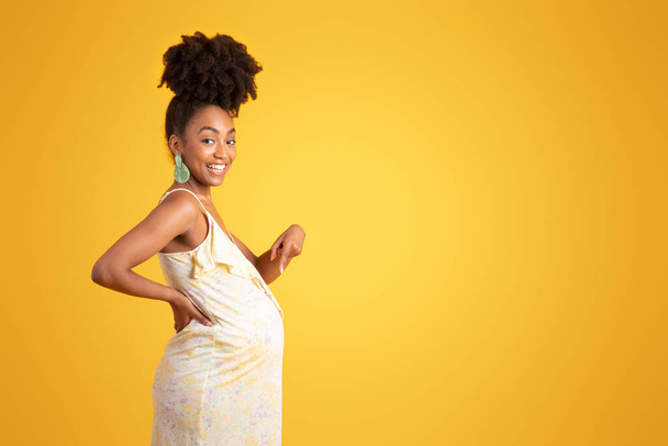 Fröhlich stolze afrikanische Millennial-Amerikanerin zeigt mit dem Finger auf dicken Bauch, isoliert auf gelbem Hintergrund, Studio. Anzeige und Angebot, Schwangerschaft, Mutterschaft, Babyerwartung und Fruchtbarkeit - Foto, Bild