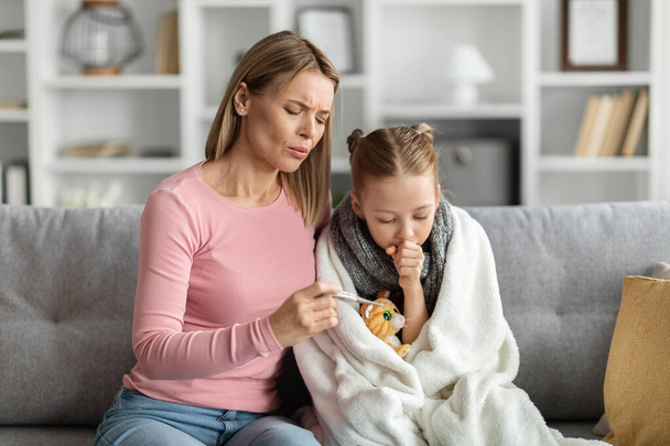 Заботливая молодая мама, проверяющая температуру своего больного ребенка дома, беспокойная мать, держащая термометр и обнимающая плохую маленькую дочь, сидящая в одеяле девочка и кашляющая в кулак - Фото, изображение