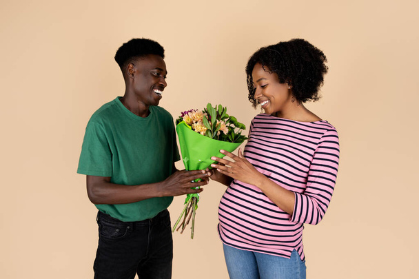 幸せな千年のアフリカ系アメリカ人男性はベージュの背景に隔離された大きな腹を持つ妻に花束を与えます。ロマンチックな関係、日付、お祝いの記念日、誕生日、子供を期待する - 写真・画像