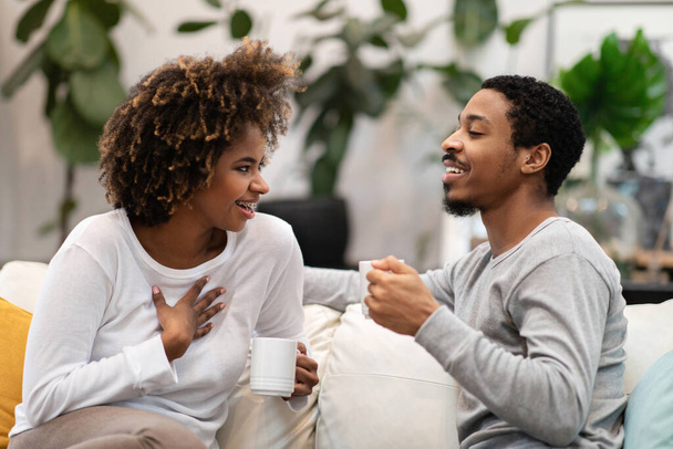 Cheerful positive loving young black spouses husband and wife wearing homewear pajamas sitting on couch in living-room, couple drinking coffee at home, have conversation and laughing - Photo, image
