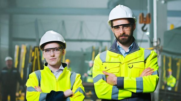Portrait of Caucasian man and woman colleagues at manufactory in hardhats, goggles and uniforms. Male and female co-workers at factory looking at camera. Team of plant production workers. - Foto, Imagen