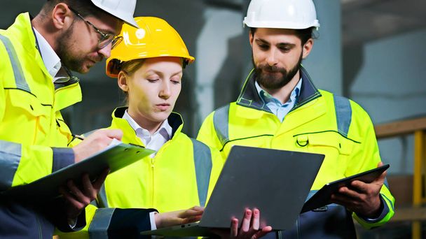 Close up of men and woman at construction site talking and discussing work with laptop computer, documents and tablet. Production plant. Males and female co-workers at building. Builders team. - Valokuva, kuva