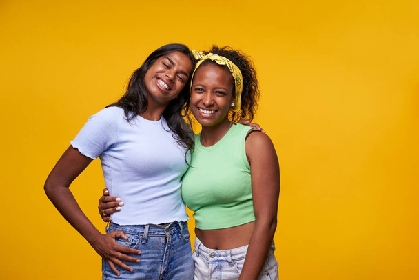 Ζευγάρι χαμογελώντας ινδική και αφρικανική αμερικανική κορίτσι θέτουν αγκαλιάζει αναζητούν χαρούμενα στην κάμερα απομονώνονται σε κίτρινο φόντο χρώμα. Ομοφυλοφιλική σχέση ευτυχισμένων λεσβιών φιλενάδων σε θετική στάση. - Φωτογραφία, εικόνα