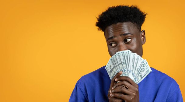 Lottery Luck. Happy young black man has just won the lottery jackpot, holding bunch of cash dollar banknotes over his face, looking at copy space, isolated on yellow studio background, banner - Photo, Image