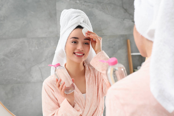 Beautiful woman in terry towel removing makeup with cotton pad near mirror indoors - 写真・画像