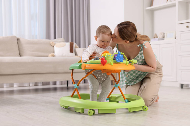 Cute boy making first steps with baby walker. Mother kissing her little son at home, space for text - Zdjęcie, obraz