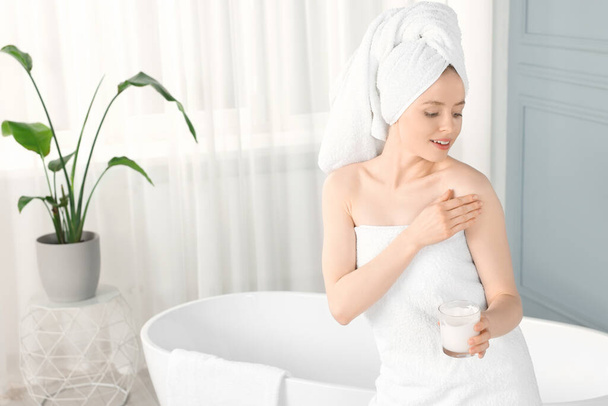 Beautiful young woman applying body cream onto shoulder in bathroom - Φωτογραφία, εικόνα