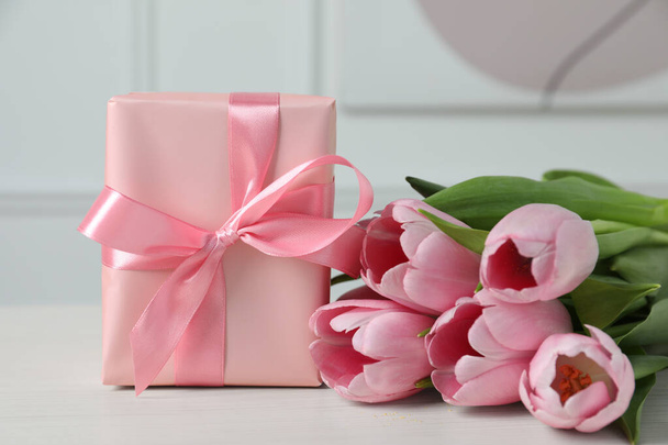 Beautiful gift box with bow and pink tulips on white table, closeup - Fotó, kép