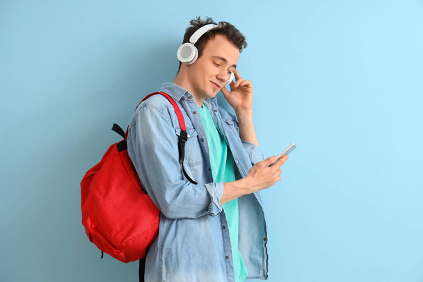 Estudiante masculino con mochila y teléfono móvil escuchando música en auriculares sobre fondo azul - Foto, imagen