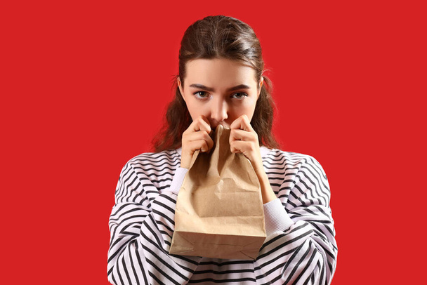 Молодая женщина с бумажным пакетом паникует на красном фоне - Фото, изображение