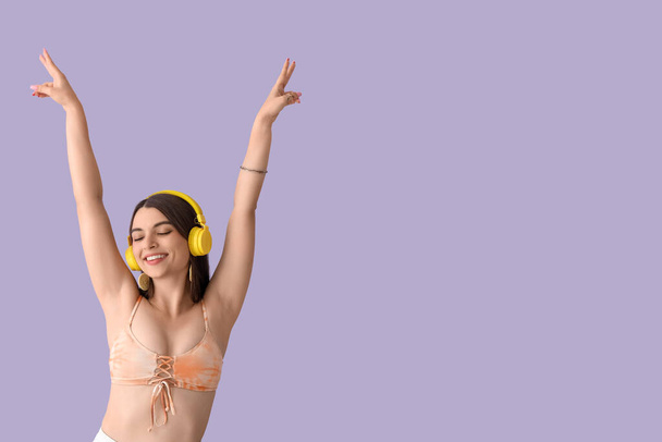Young woman in headphones showing victory gesture on lilac background - Photo, Image
