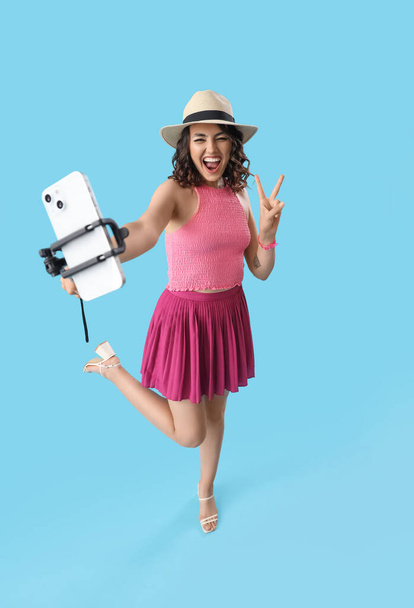 Щаслива молода жінка з мобільним телефоном бере селфі на синьому фоні - Фото, зображення