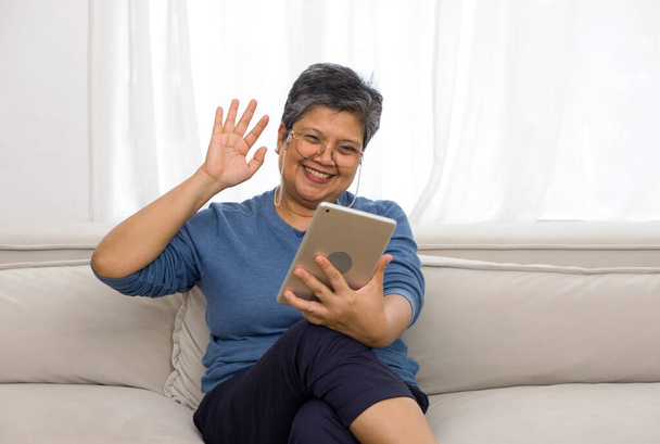 Aasian harmaat hiukset vanha nainen heiluttaa kättä etäviestinnän kautta tabletin tietokoneen monitorin istuessa sohvalla olohuoneessa. - Valokuva, kuva