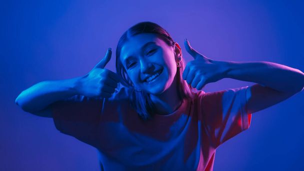 Like gesture. Cool offer. Positive recommendation. Happy smiling woman in vibrant neon light showing thumbs up on blue background. - 写真・画像