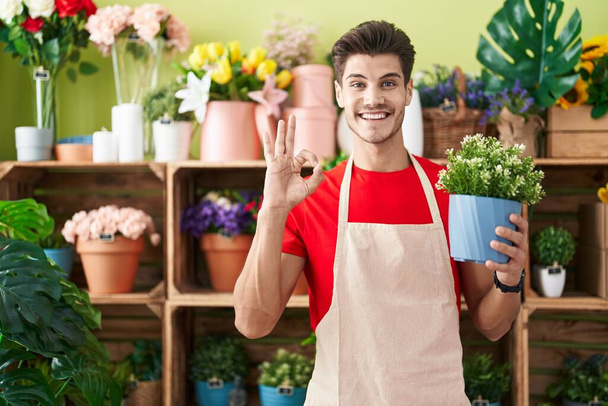 Young hispanic man working at florist shop holding plant doing ok sign with fingers, smiling friendly gesturing excellent symbol  - Fotografie, Obrázek