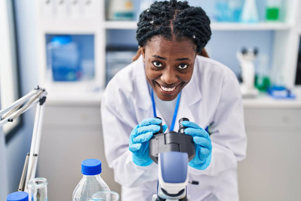 Африканська американська жінка - науковець, яка впевнено посміхається за допомогою мікроскопа в лабораторії. - Фото, зображення