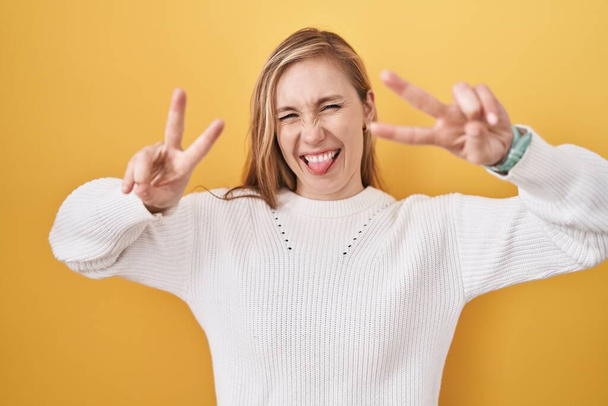 Fiatal fehér nő fehér pulóvert visel sárga háttér felett mosolyogva, nyelvvel kifelé mutatva mindkét kezét, amint győzelmi jelet csinál. Második..  - Fotó, kép