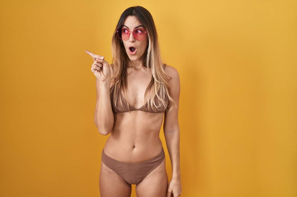 Sarı arka planda bikini giyen genç İspanyol kadın parmaklarını yana çevirerek şaşırdı, ağzı açık hayret içinde kaldı..  - Fotoğraf, Görsel