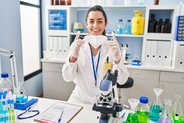 Νεαρή όμορφη ισπανίδα επιστήμονας χαμογελώντας αυτοπεποίθηση κρατώντας ιατρική μάσκα στο εργαστήριο - Φωτογραφία, εικόνα