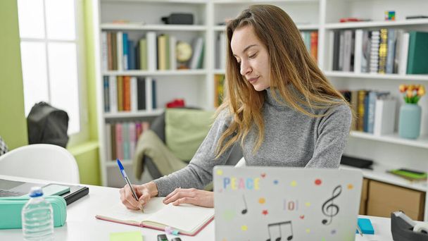 Young caucasian woman student using laptop writing on notebook at library university - Valokuva, kuva