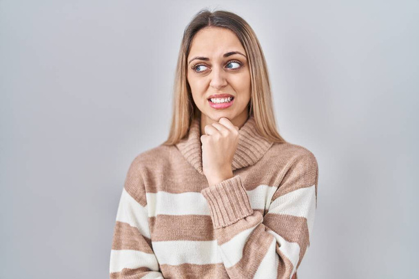 Giovane donna bionda che indossa maglione collo alto su sfondo isolato pensando preoccupato per una domanda, preoccupato e nervoso con mano sul mento  - Foto, immagini