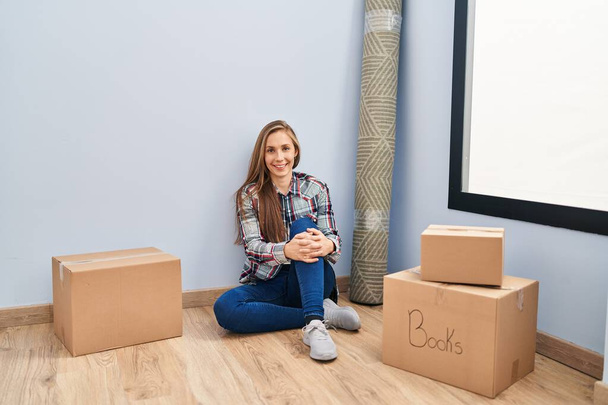 Young blonde woman sitting on the floor moving to a new home with a happy and cool smile on face. lucky person.  - Photo, image