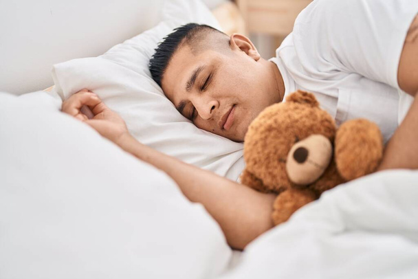 Молодой латиноамериканец обнимает плюшевого мишку, лежащего на кровати и спит в спальне - Фото, изображение