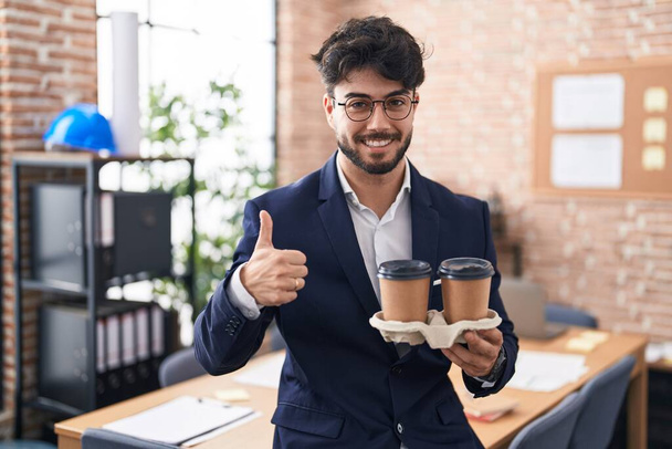Hombre hispano con barba trabajando en la oficina sosteniendo café sonriendo feliz y positivo, pulgar hacia arriba haciendo excelente y signo de aprobación  - Foto, imagen