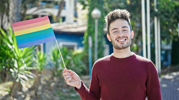 Junger hispanischer Mann lächelt selbstbewusst mit Regenbogenfahne im Park - Foto, Bild