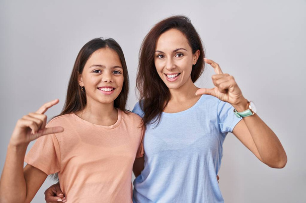 Young mother and daughter standing over white background smiling and confident gesturing with hand doing small size sign with fingers looking and the camera. measure concept.  - Valokuva, kuva