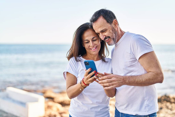 Мужчина и женщина среднего возраста используют смартфон, стоящий вместе на берегу моря - Фото, изображение